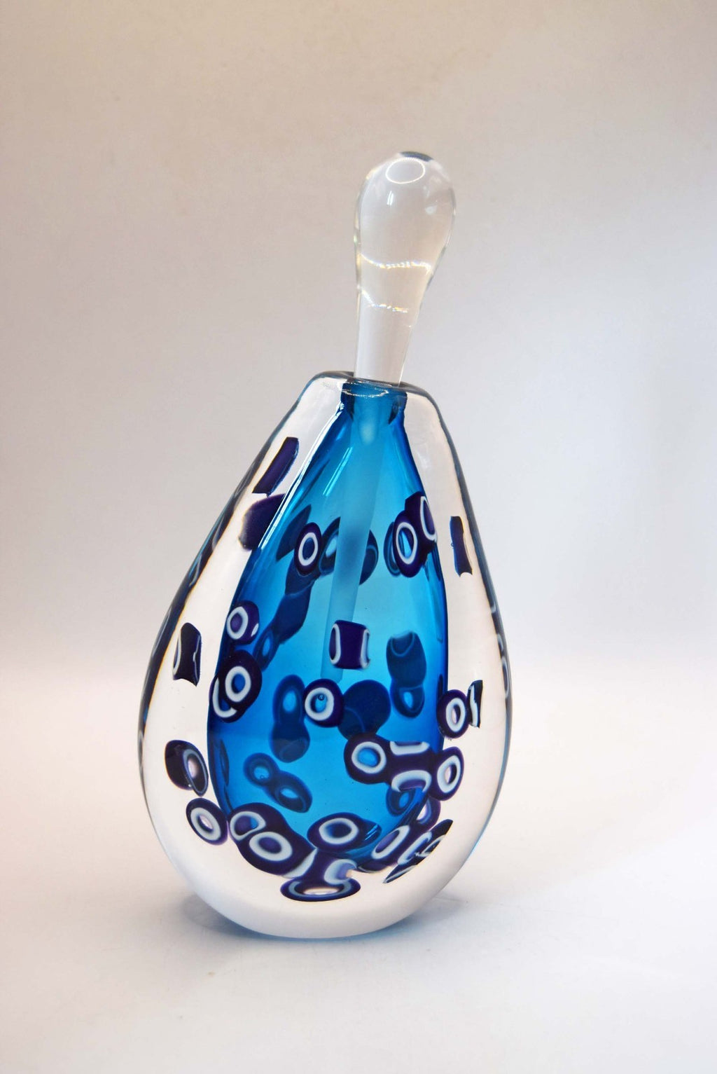 A hand-blown glass vase with a teardrop shape, featuring a vibrant blue center filled with dark blue and white circular patterns, a white neck, and a white base, set against a plain light background.