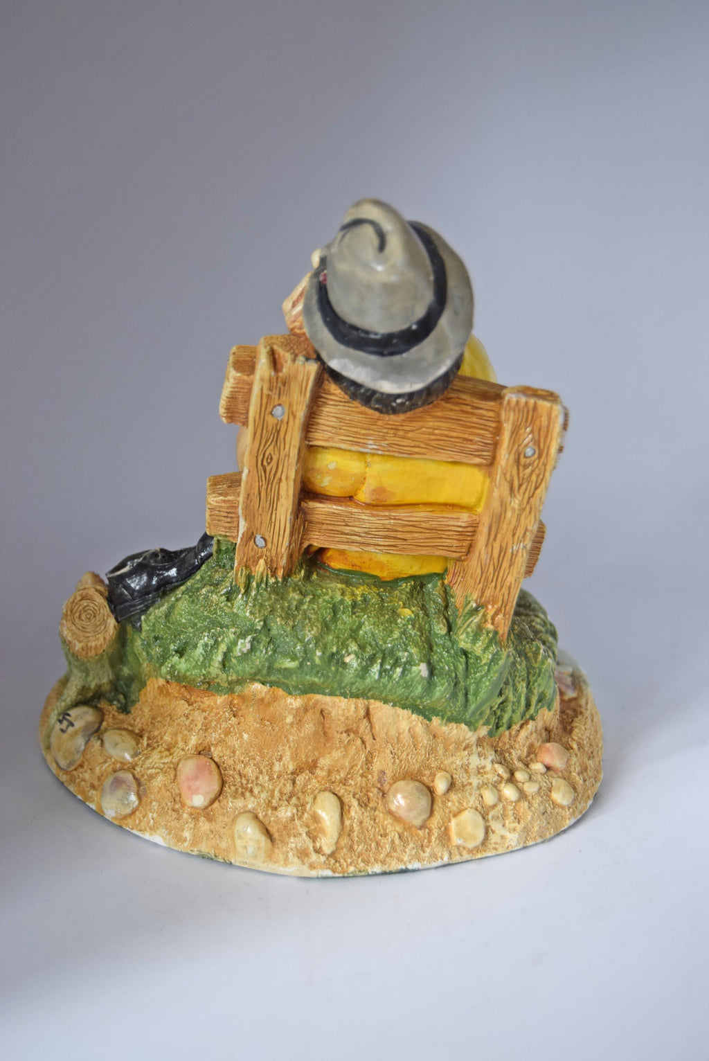 Decorative figurine of a person sitting on a wooden bench with a hat on their head, on a textured base.