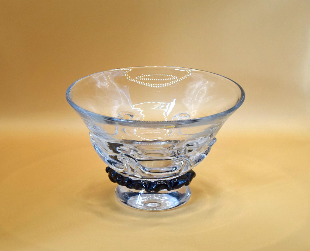 A clear glass bowl with a flared rim and pedestal base, featuring a dark blue beaded band around the lower middle, set against a warm beige background.