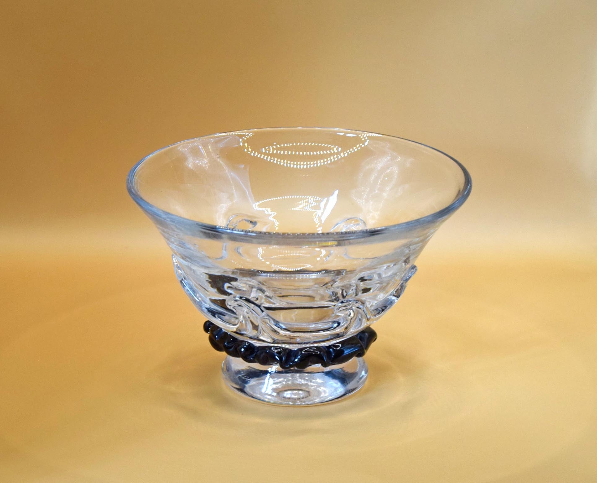 A clear glass bowl with a flared rim and pedestal base, featuring a dark blue beaded band around the lower middle, set against a warm beige background.