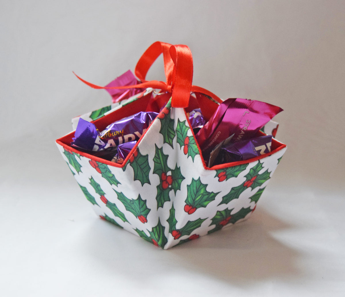 A small decorative gift basket or candy box wrapped in white paper with a green holly and red berry pattern, trimmed with red edging and topped with a red ribbon handle. The basket is filled with wrapped chocolates on a plain light background.