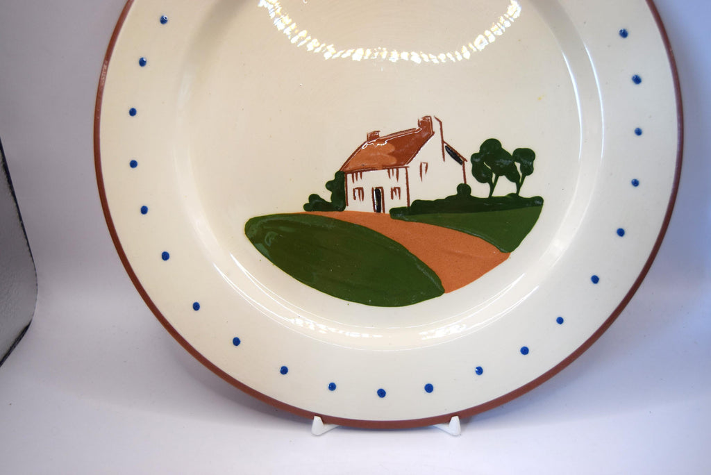 A vintage ceramic plate with a hand-painted country scene in the center, featuring a white house, green hills, and an orange path. The plate has a cream-colored glaze, a brown rim, and blue polka dots around the edge, displayed on a white surface.