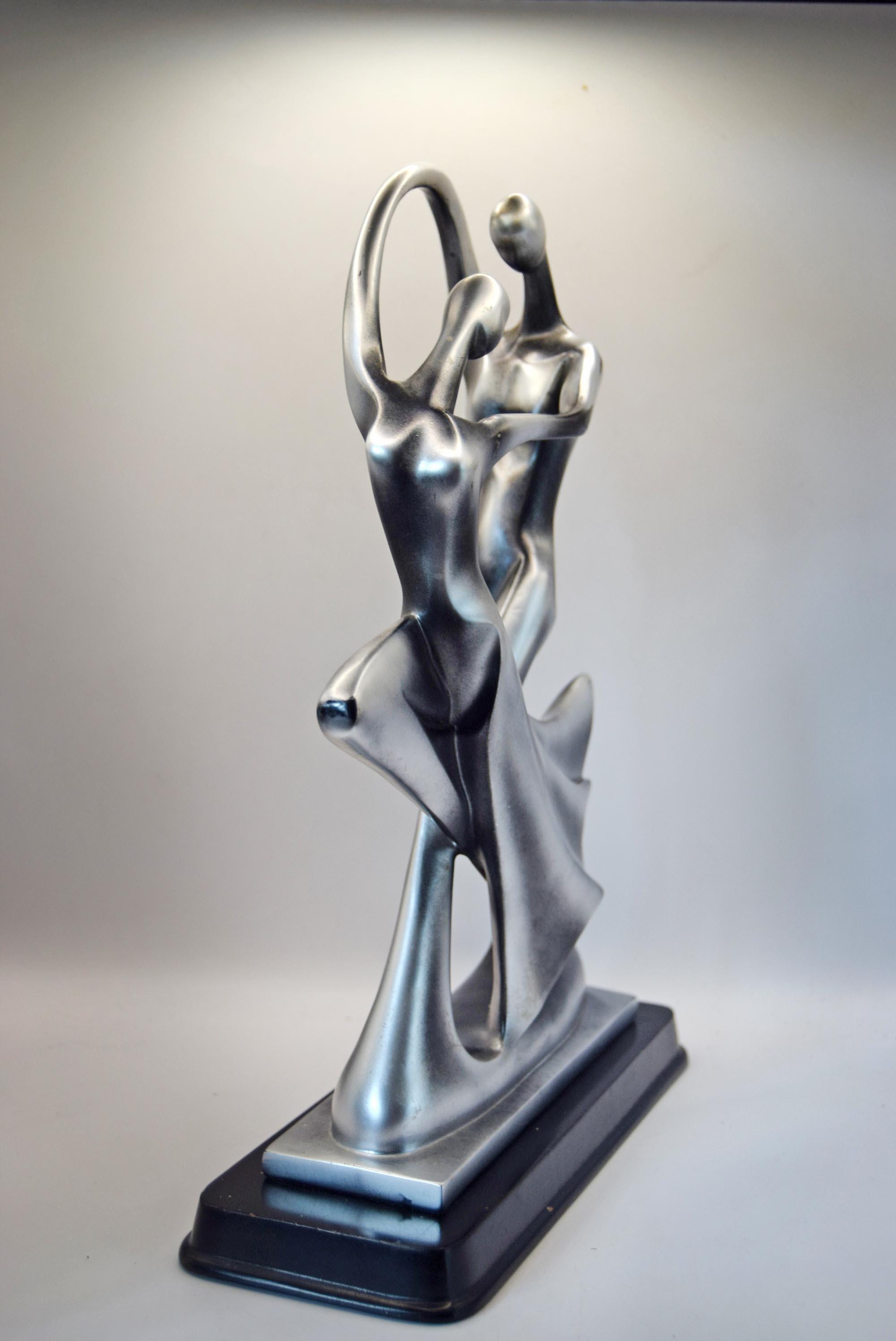 A modern abstract sculpture of a dancing couple in metallic silver, mounted on a tiered black base against a neutral gray studio background.
