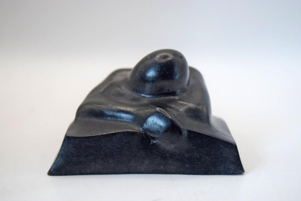 A small black sculptural object with a smooth, rounded organic top and a matte base, photographed against a plain light background.