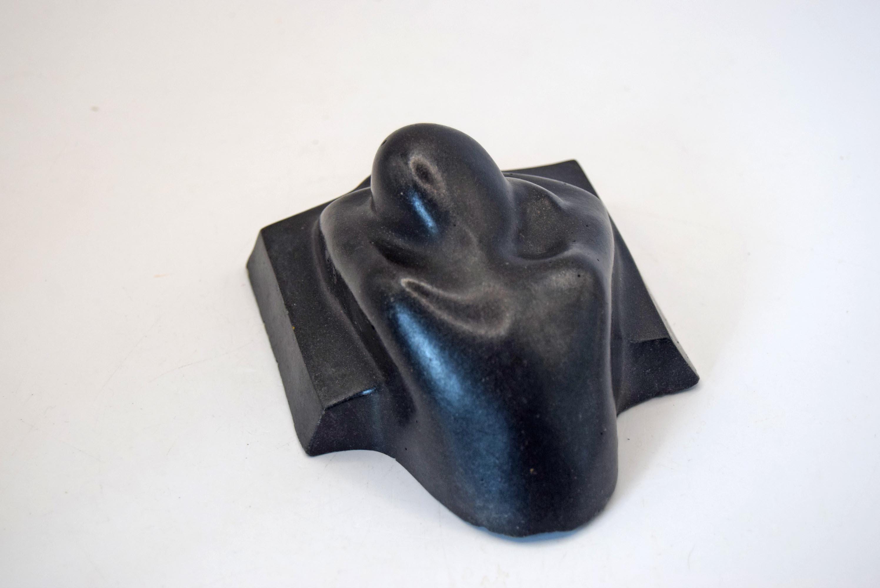 A black sculptural object with a smooth, organic, abstract form mounted on a square base, photographed against a plain white background.