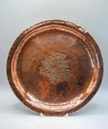 A hammered copper serving platter with a central floral engraving, displayed on a small stand against a neutral gray background.