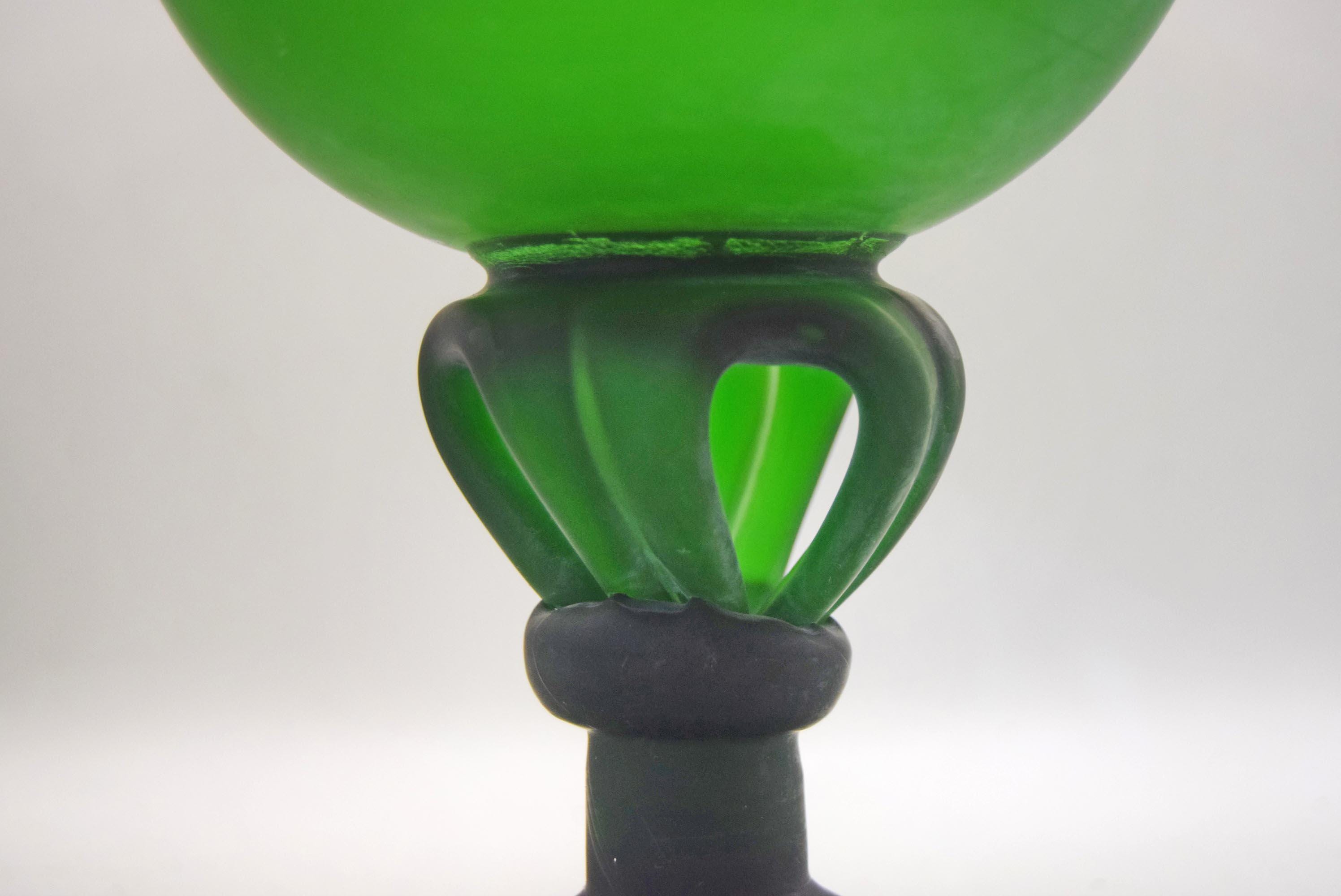 A close-up of a green glass goblet or cup with a dark gray stem and a flared, ribbed base, set against a neutral gray background.