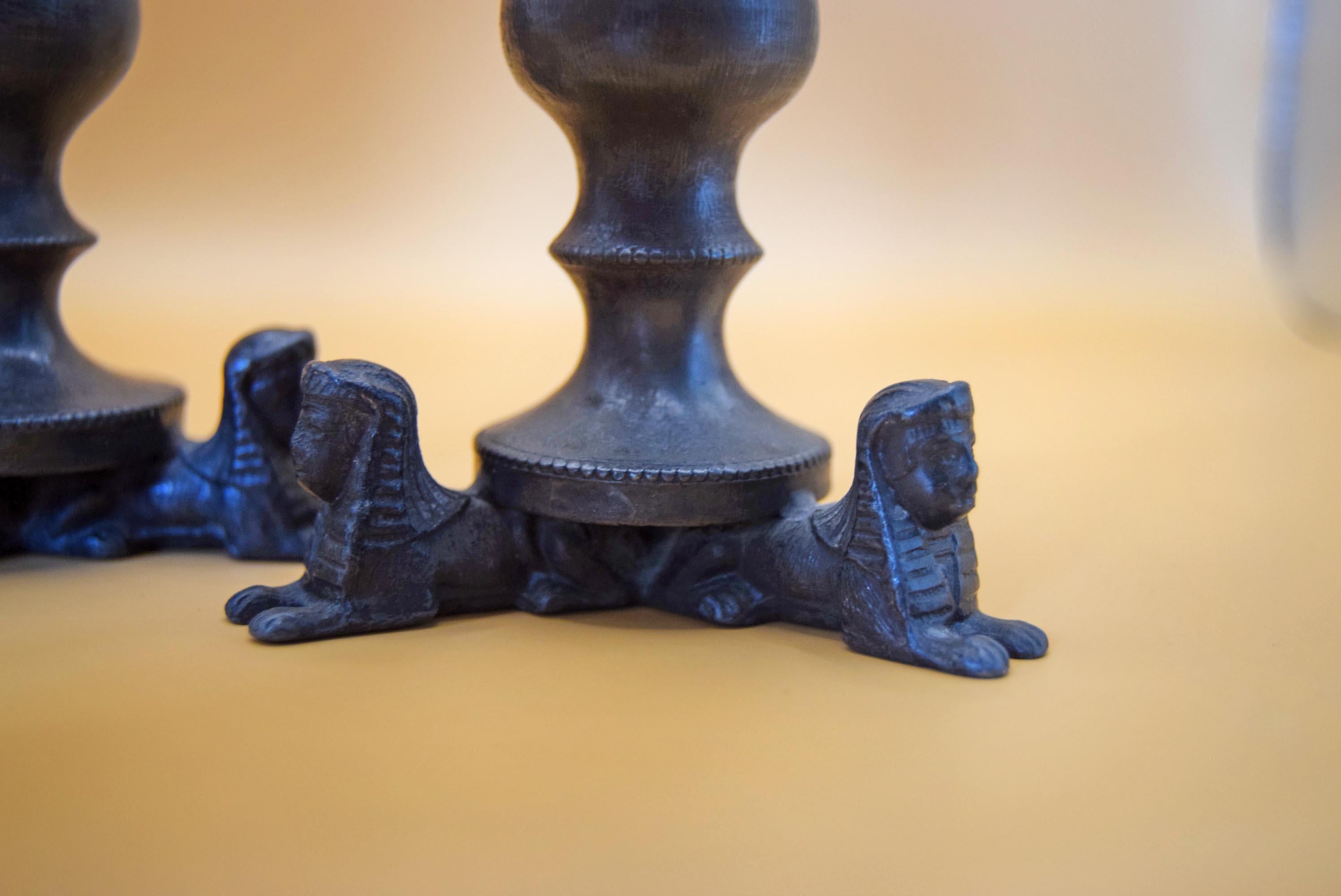 Close-up of a dark metal decorative object with sphinx figures supporting the base, shown on a warm beige surface with a softly blurred background.
