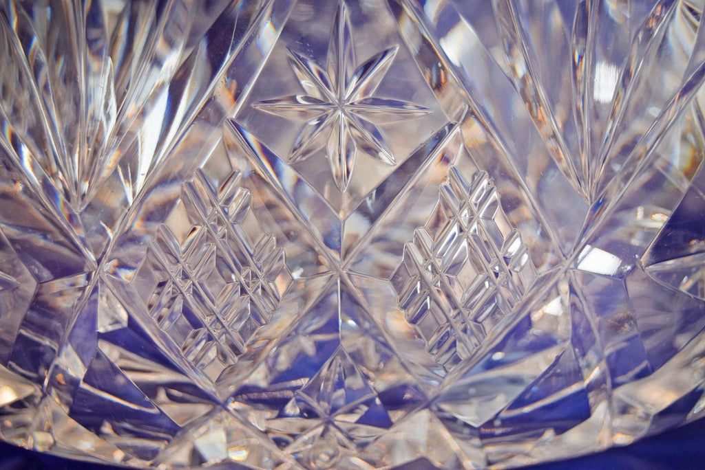 Heavy cut glass fruit bowl, deeply cut geometric motifs, combining radiant hobstars, fanned wheel cuts, and diamond cross-hatching that catch the light beautifully from every angle. It is topped with a dramatic, flared scalloped rim detailed with sharp sawtooth edging.