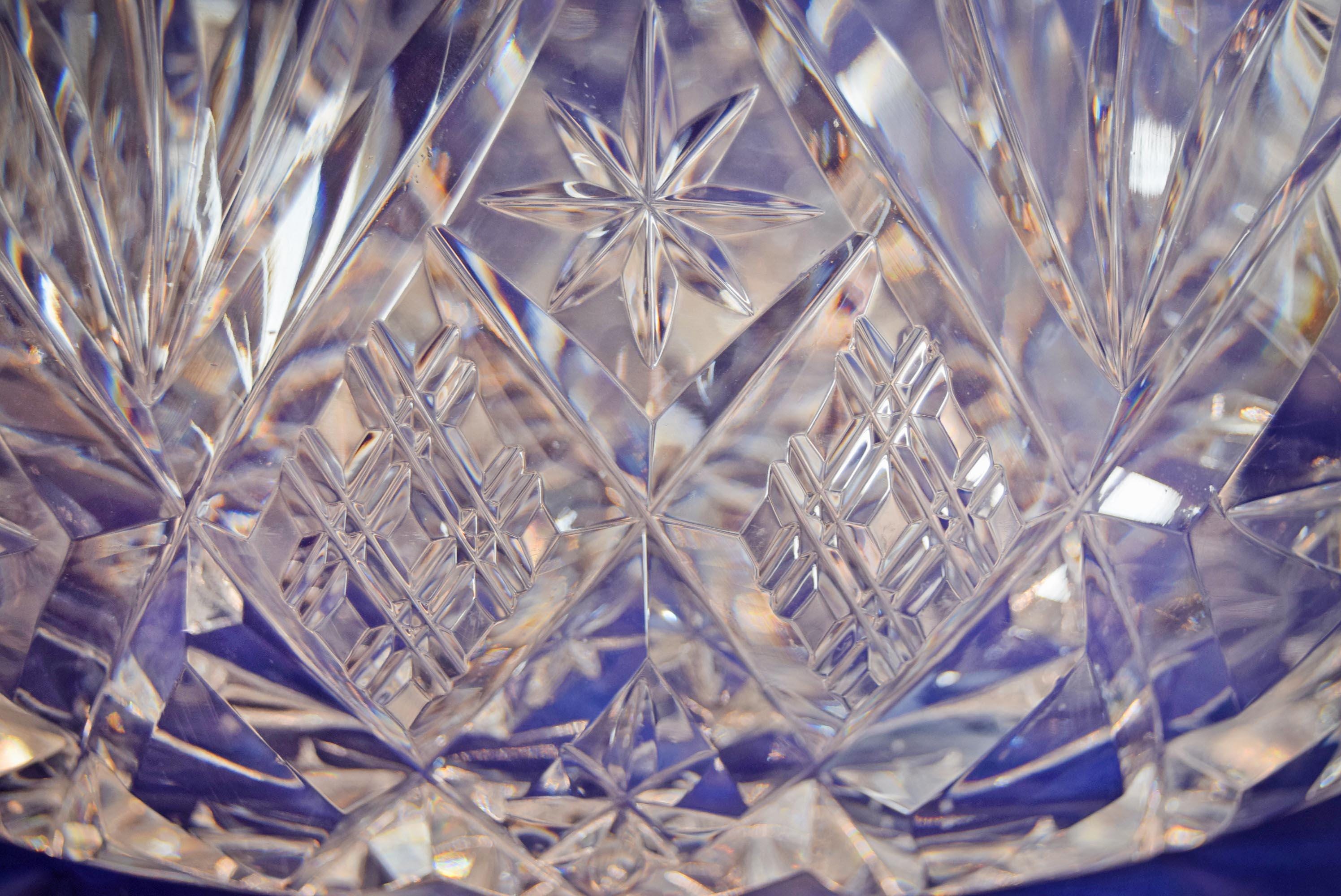 Heavy cut glass fruit bowl, deeply cut geometric motifs, combining radiant hobstars, fanned wheel cuts, and diamond cross-hatching that catch the light beautifully from every angle. It is topped with a dramatic, flared scalloped rim detailed with sharp sawtooth edging.