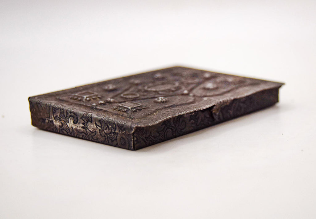 Late-Victorian to early 20th-century antique pocket box disguised as a miniature book. Originally manufactured between 1880 and 1920, this charming novelty case was intended for carrying powdered snuff tobacco or medicinal pills. It features an exceptionally detailed, machine-stamped exterior that beautifully mimics the elaborate repoussé and chased metalwork of traditional 17th-century European book covers.