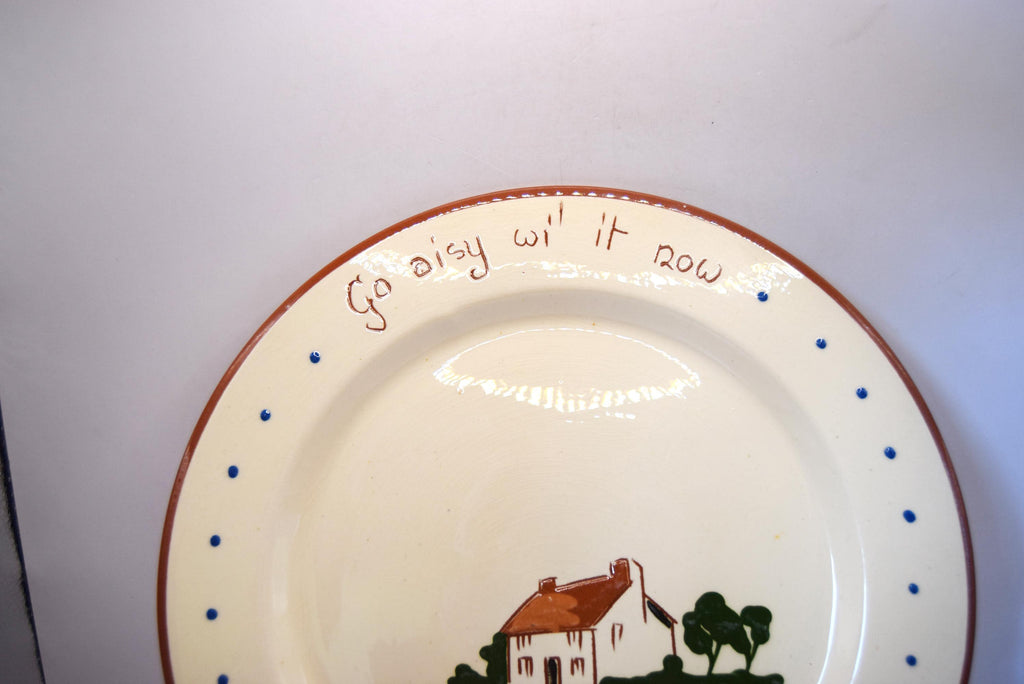 A close-up of a ceramic plate with a cream-colored glaze, featuring the handwritten phrase 'Go Daisy Wit' Her' around the rim, decorated with blue dots and a brown border. The bottom of the plate shows a small painted illustration of a house and trees.