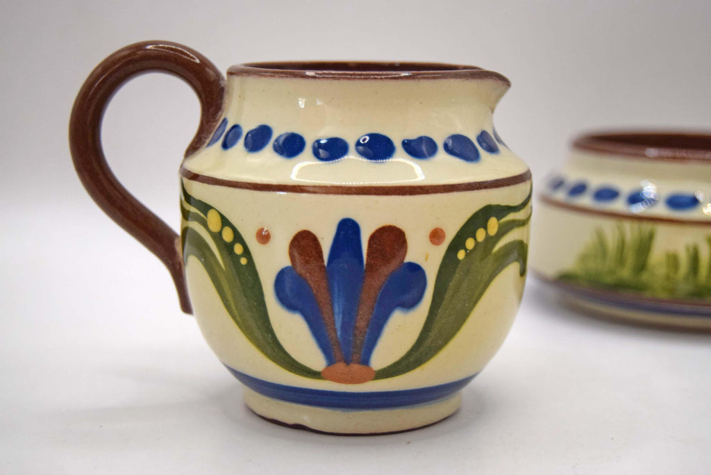 A small ceramic pitcher with a brown handle and rim, decorated with a cream-colored glaze, blue dots, green leaves, and a blue and brown floral motif. A second matching pitcher is partially visible in the background on a white surface.