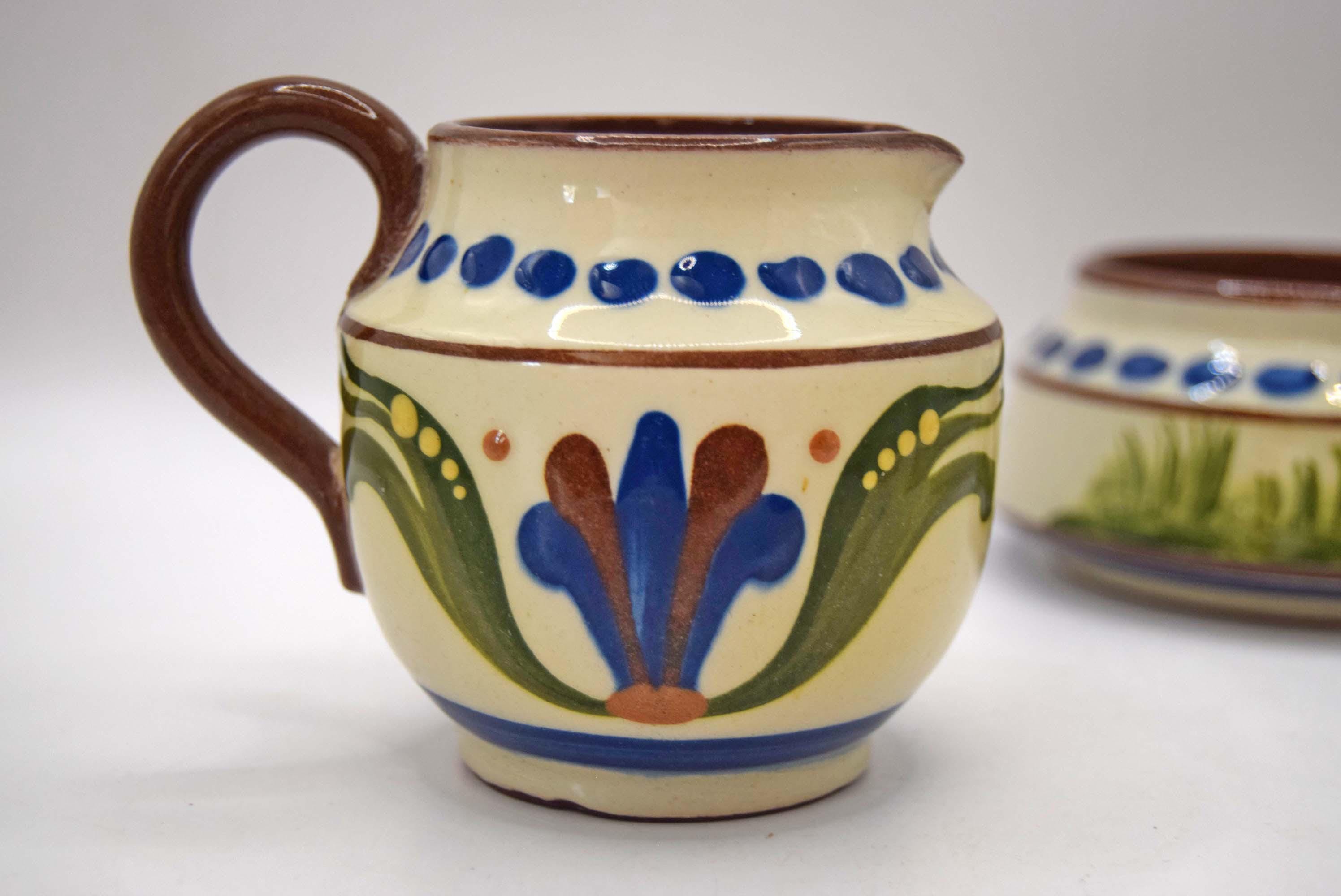 A small ceramic pitcher with a brown handle and rim, decorated with a cream-colored glaze, blue dots, green leaves, and a blue and brown floral motif. A second matching pitcher is partially visible in the background on a white surface.