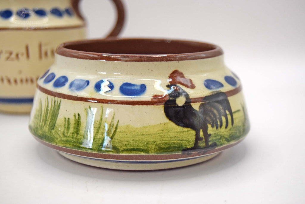 A small handmade ceramic bowl with a hand-painted rooster and green grass design, featuring blue polka dots and a brown rim, shown on a white background.