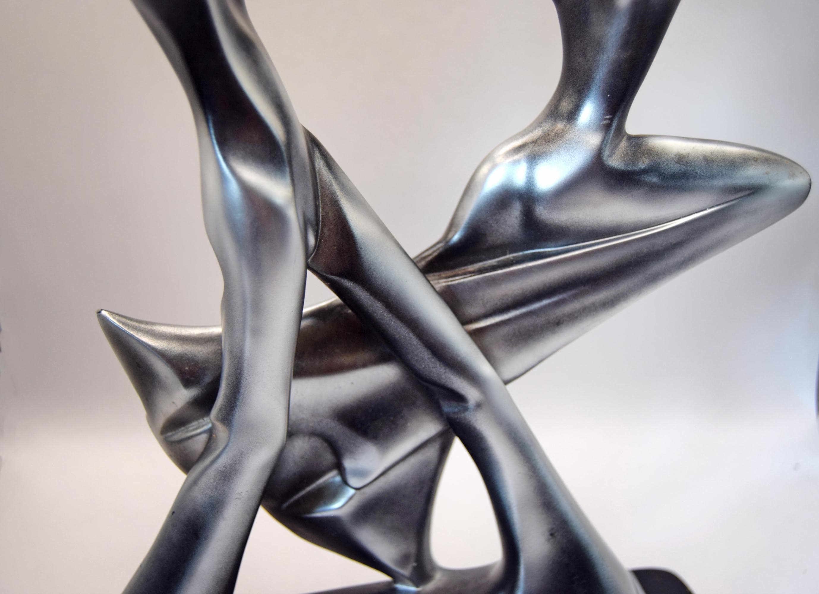 Abstract metallic sculpture with flowing, intertwined organic forms in a brushed silver finish, photographed against a soft light gray background.