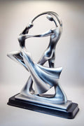 A modern abstract sculpture of two dancing figures in metallic silver with a black rectangular base, photographed against a neutral light gray background.