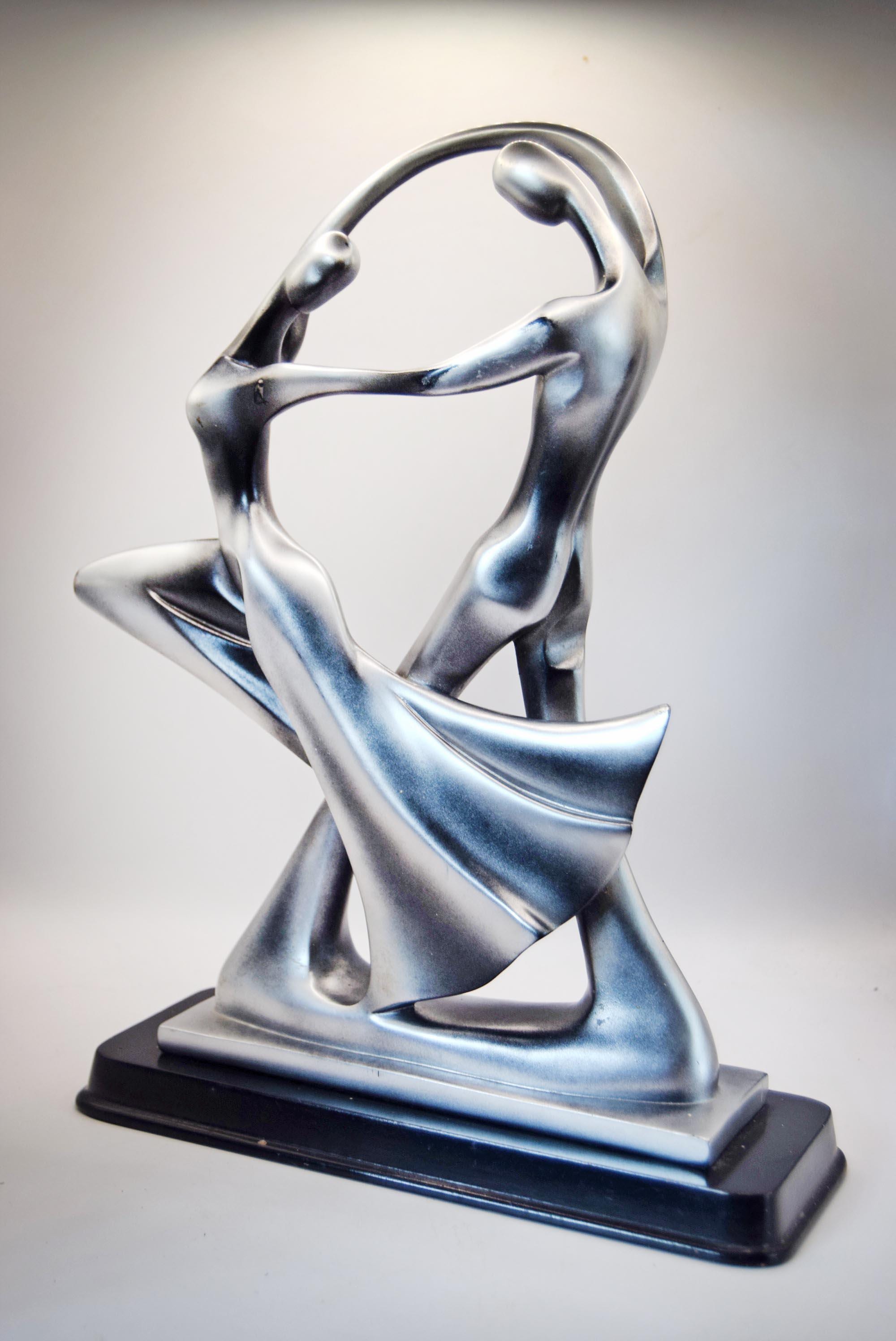A modern abstract sculpture of two dancing figures in metallic silver with a black rectangular base, photographed against a neutral light gray background.