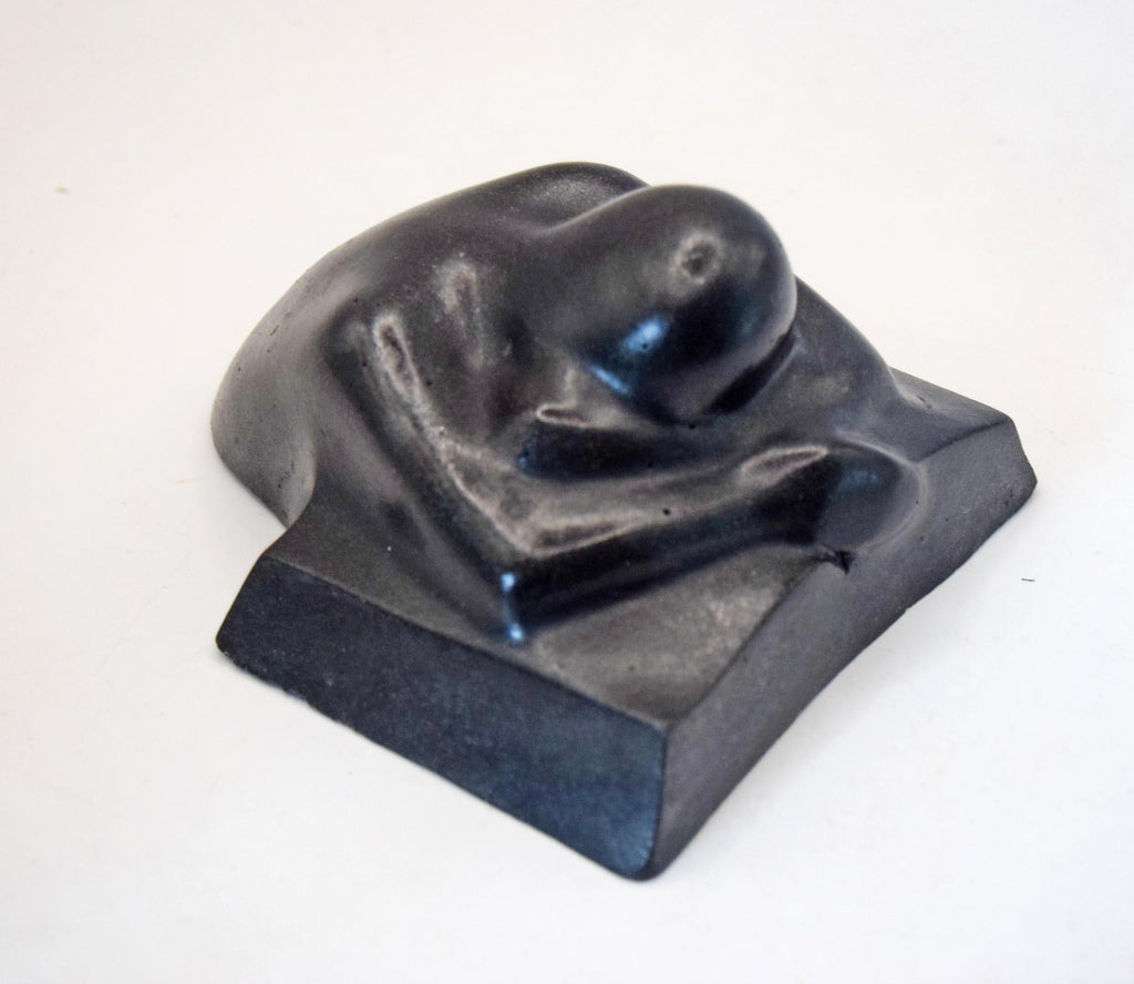 A black sculptural figurine of a curled-up human figure on a rectangular base, photographed against a plain light background.