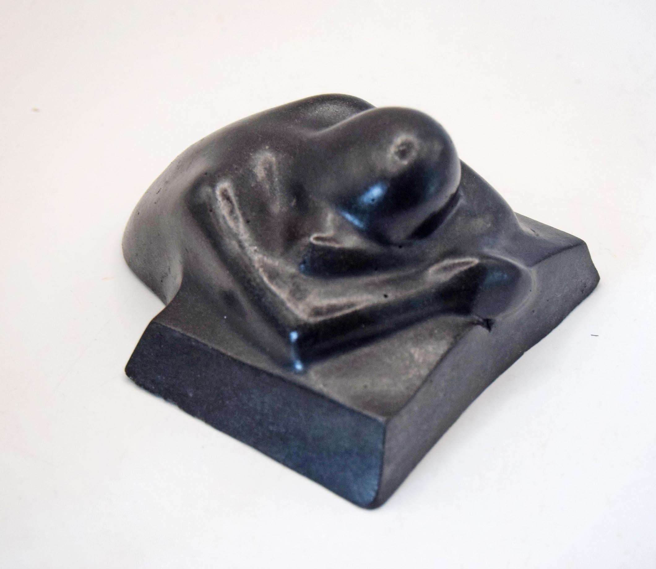 A black sculptural figurine of a curled-up human figure on a rectangular base, photographed against a plain light background.