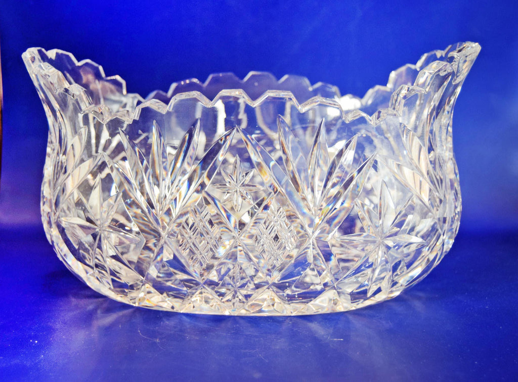 Heavy cut glass fruit bowl, deeply cut geometric motifs, combining radiant hobstars, fanned wheel cuts, and diamond cross-hatching that catch the light beautifully from every angle. It is topped with a dramatic, flared scalloped rim detailed with sharp sawtooth edging.