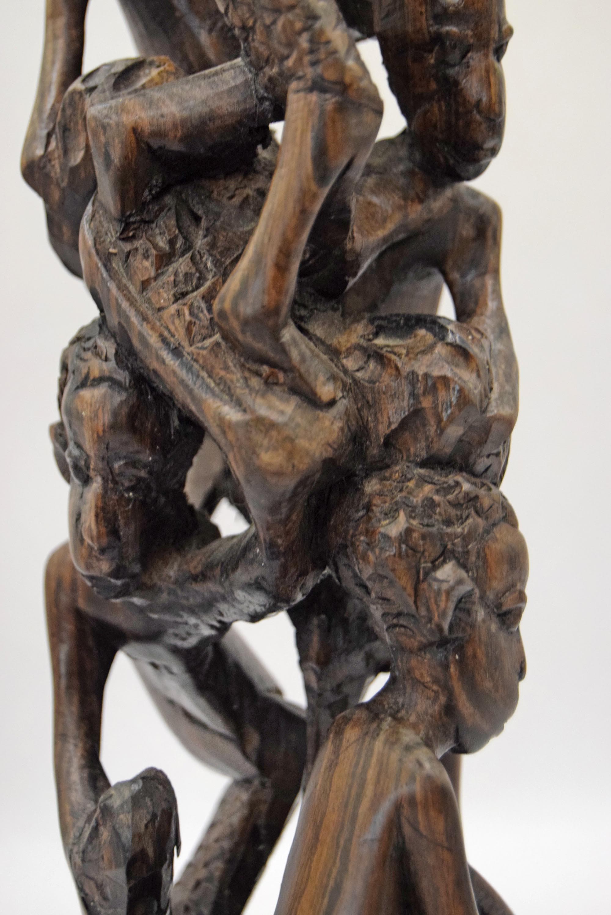 A close-up of a dark brown carved wooden sculpture depicting multiple human figures in an intertwined, dynamic composition against a plain light background.