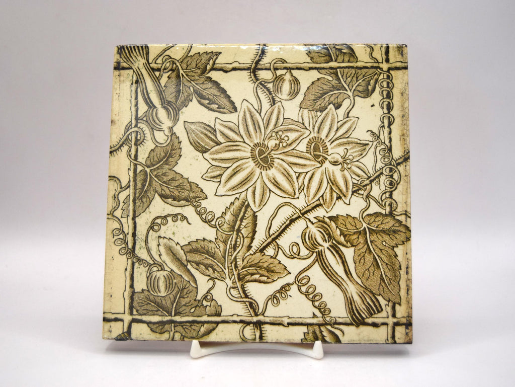 A square ceramic tile with a vintage botanical pattern of passion flowers and leaves in olive green on a cream background, displayed on a small white stand against a neutral gray background.