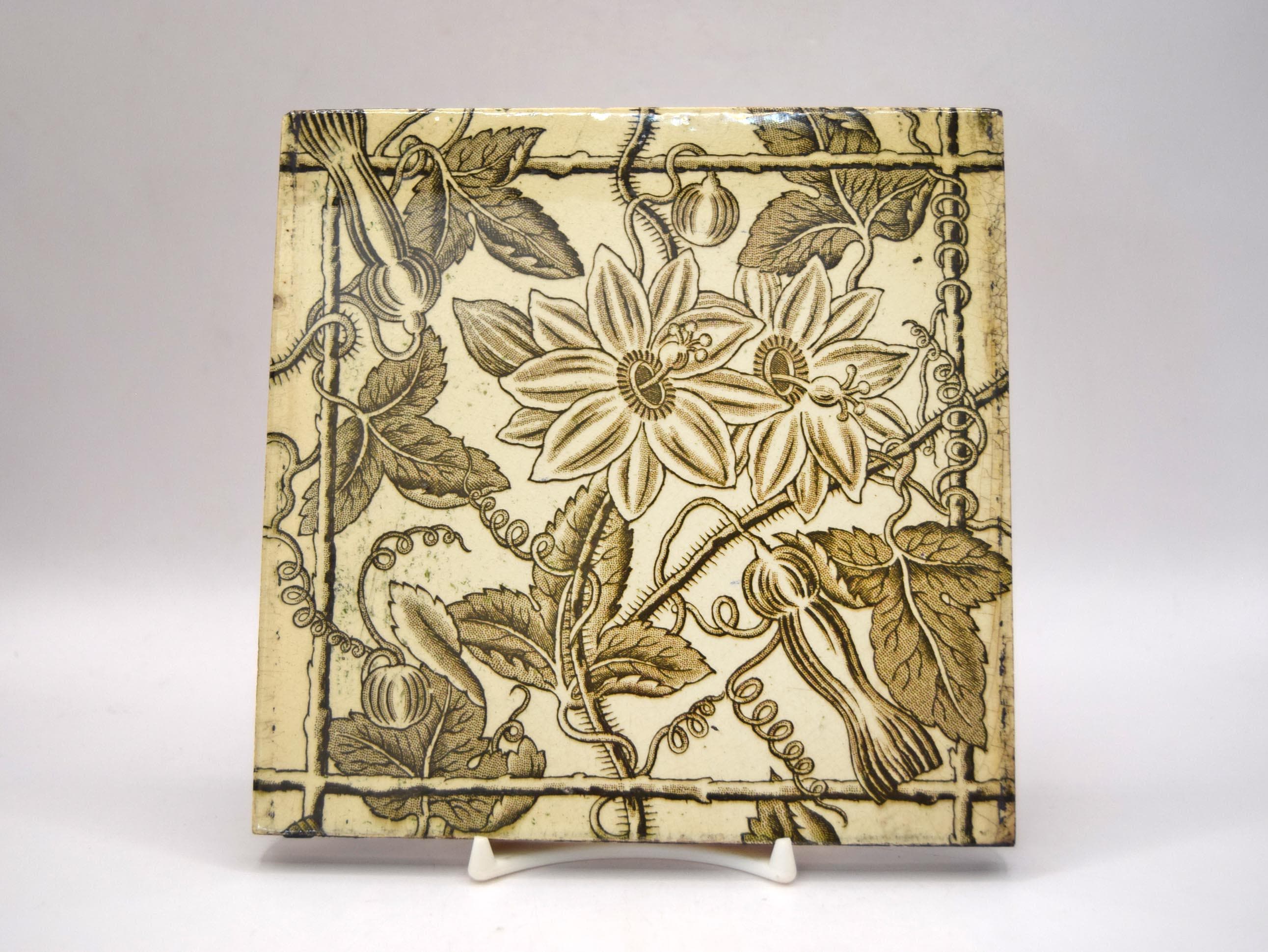 A square ceramic tile with a vintage botanical pattern of passion flowers and leaves in olive green on a cream background, displayed on a small white stand against a neutral gray background.