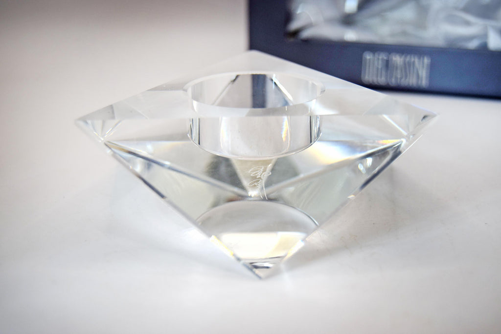 A clear faceted crystal candle holder with a diamond-like geometric design and a central circular cup, shown on a light surface with a blurred dark blue box in the background.