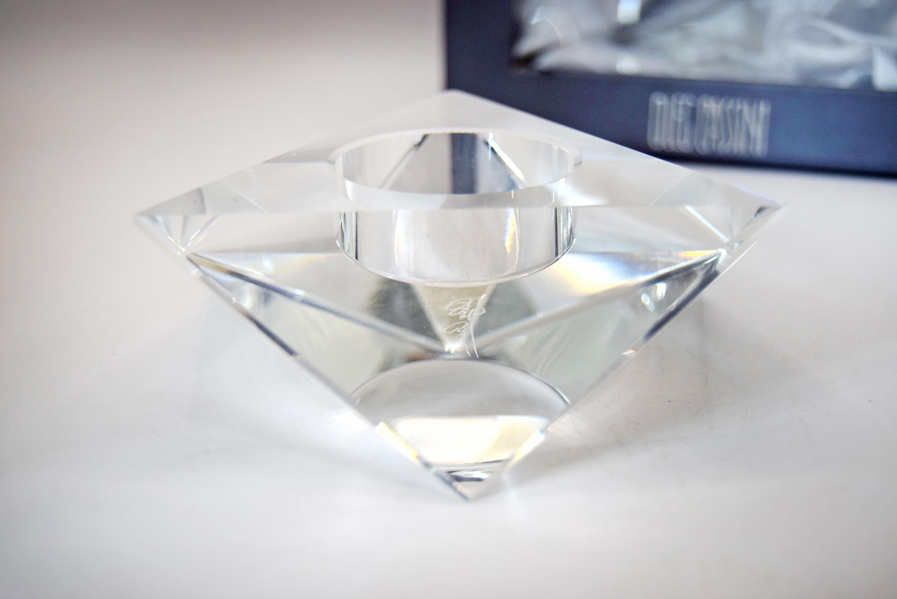 A clear faceted crystal candle holder with a diamond-like geometric design and a central circular cup, shown on a light surface with a blurred dark blue box in the background.