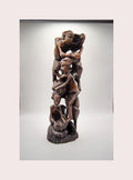 A dark brown carved wooden sculpture depicting a stacked, intertwined human figure form, displayed against a neutral light gray background.