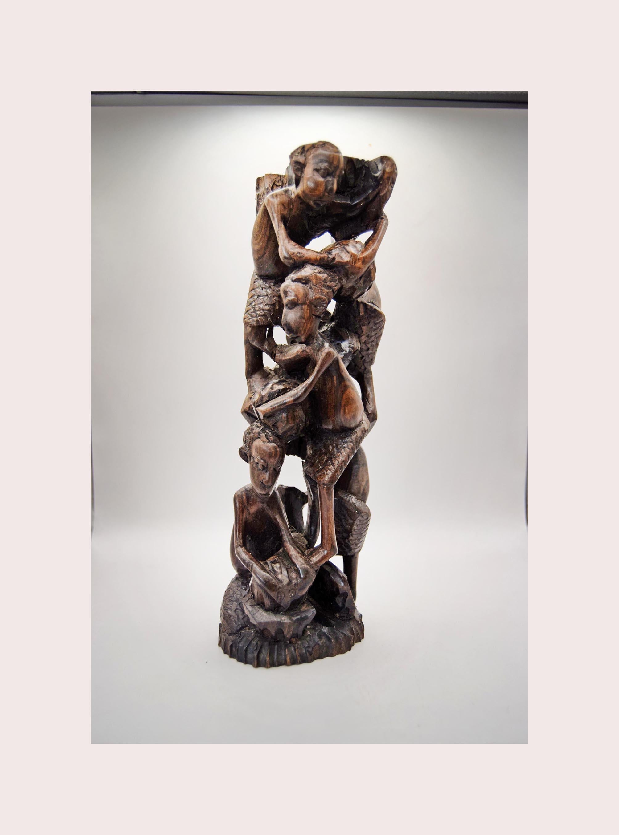 A dark brown carved wooden sculpture depicting a stacked, intertwined human figure form, displayed against a neutral light gray background.