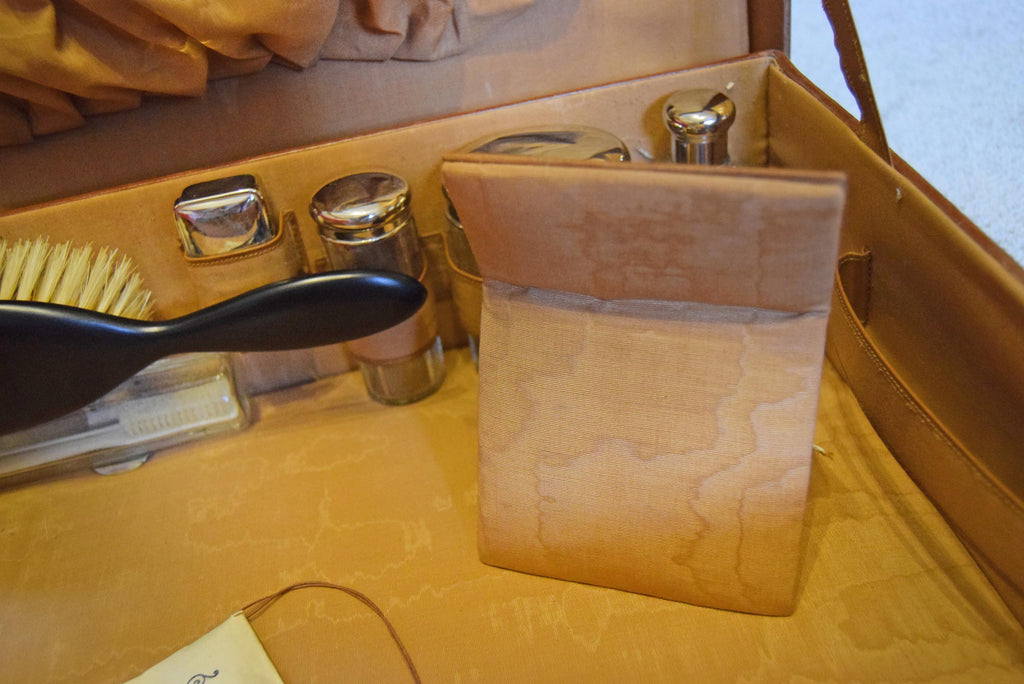 A close-up of a tan leather or faux-leather accessory case or organizer, showing a rectangular fabric pouch or cover standing inside a compartment alongside small metallic containers and a black-handled brush.