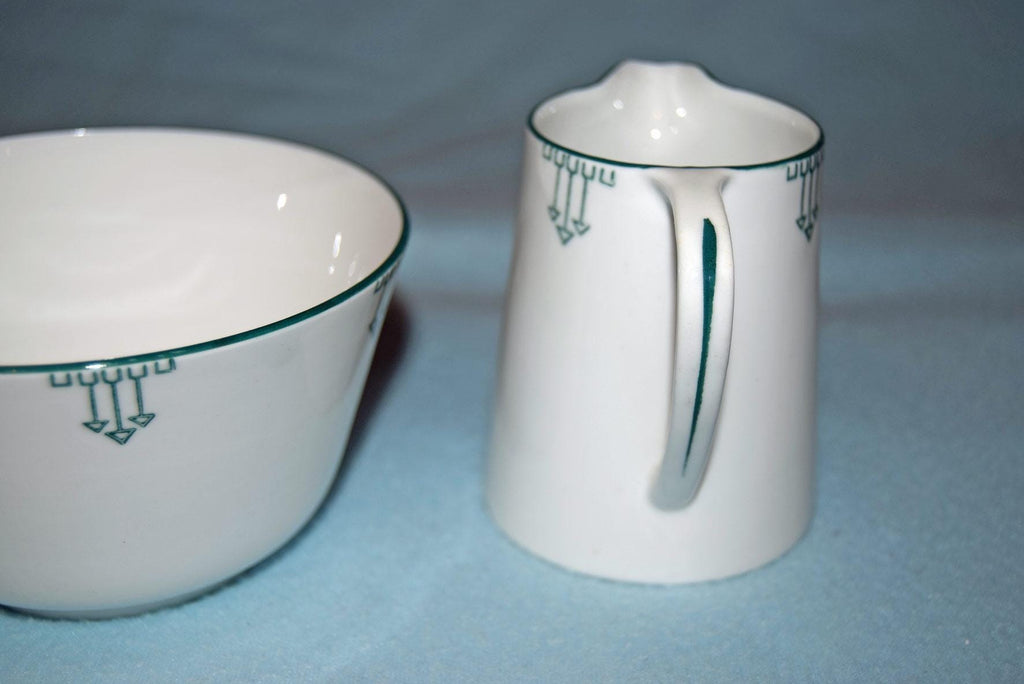 White ceramic bowl and matching pitcher with a thin green geometric border pattern, set against a soft blue background.