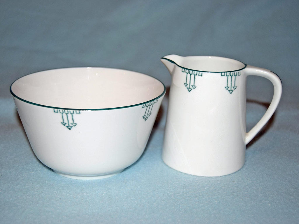 A white ceramic bowl and matching creamer pitcher with a green geometric pattern, set against a light blue background.