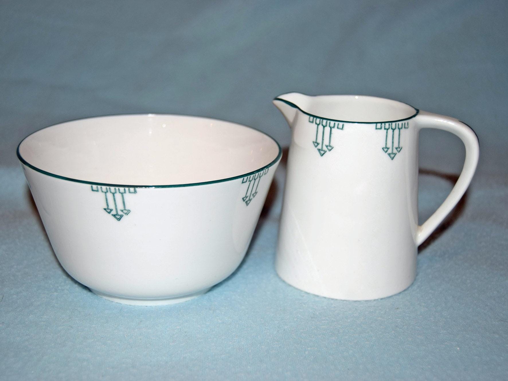 A white ceramic bowl and matching creamer pitcher with a green geometric pattern, set against a light blue background.