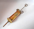 A vintage brass syringe-style pump with a circular medallion on the barrel, photographed diagonally on a plain off-white background.