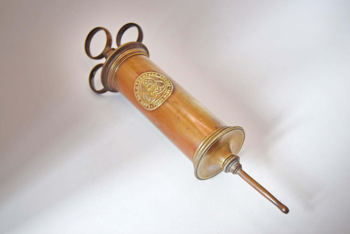A vintage brass syringe-style hand pump with a circular medallion on the side, photographed diagonally against a white studio background.