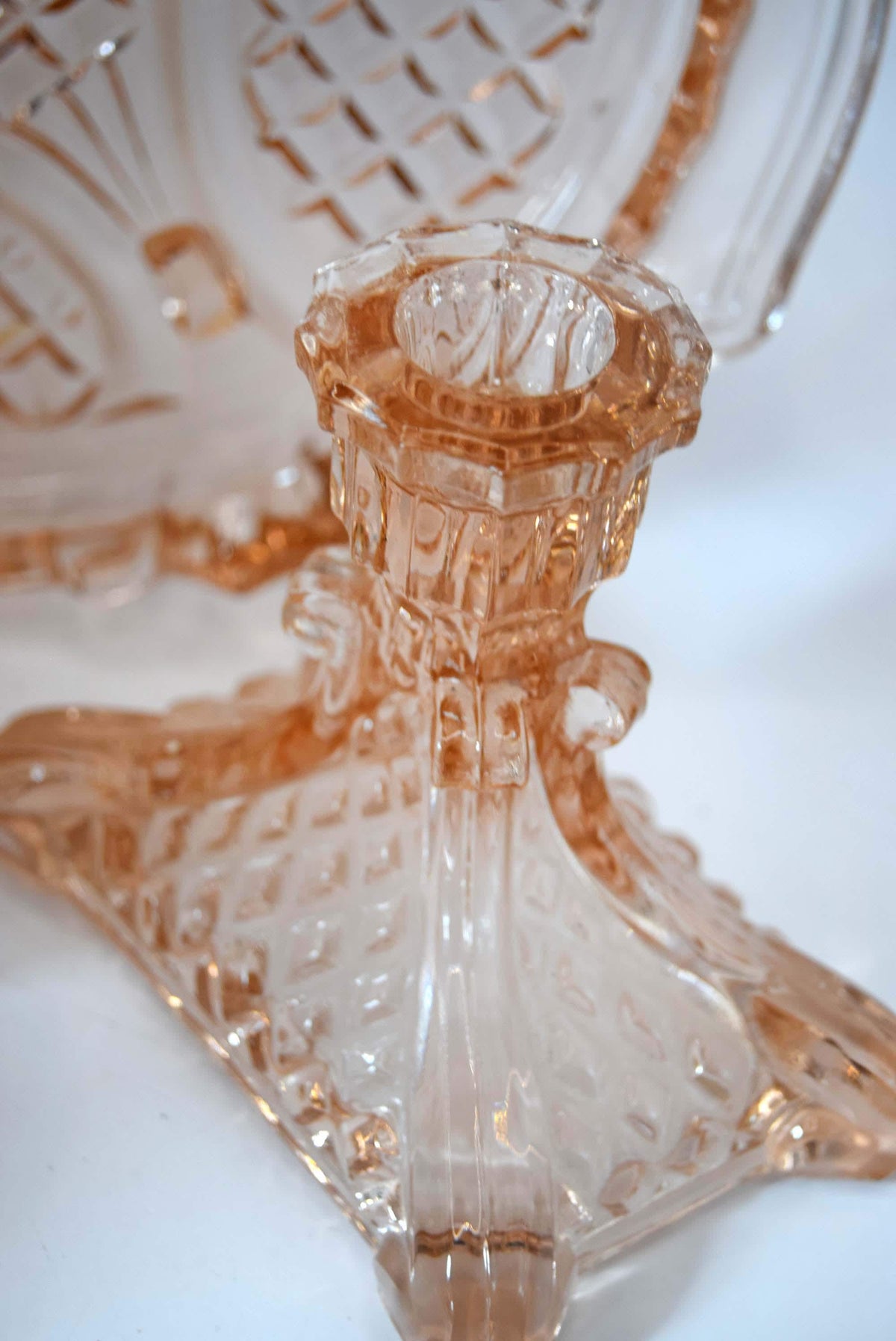 A close-up of a clear, pale pink cut-glass candlestick holder with ornate geometric and diamond-patterned detailing, set against a softly blurred white and pink background.