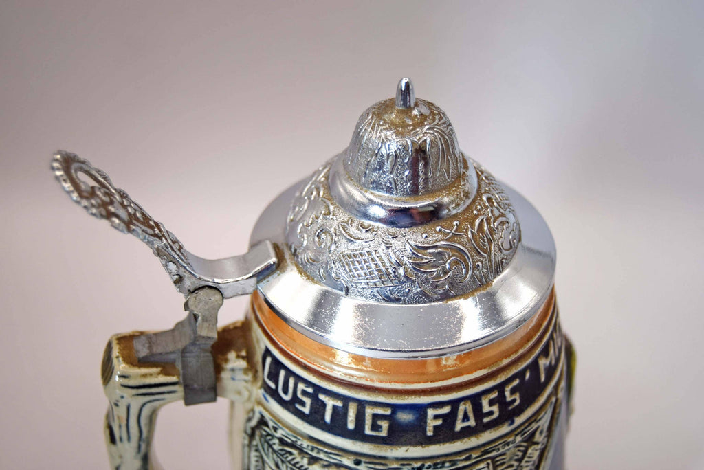 Close-up of a traditional German beer stein with a polished silver pewter lid, ornate scrollwork, and a dark blue band with white lettering that reads 'LUSTIG FASS MANN'.