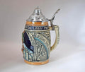 A traditional German ceramic beer stein with a pewter lid and handle, featuring relief carvings of a knight in armor and blue and orange glazes, set against a plain light gray background.