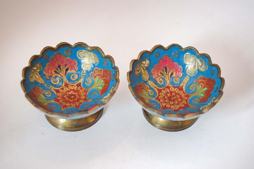 vintage solid brass pedestal bowls featuring vibrant hand-painted Champlevé-style enamel work.
The interior and exterior of each bowl showcase an intricate floral and leaf pattern set against a brilliant turquoise-blue enamel background. The design is framed by a distinct, hand-etched scalloped rim and elevated on a classic flared brass base.