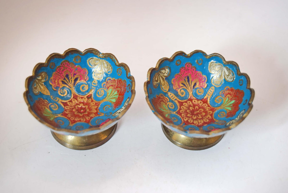 vintage solid brass pedestal bowls featuring vibrant hand-painted Champlevé-style enamel work.
The interior and exterior of each bowl showcase an intricate floral and leaf pattern set against a brilliant turquoise-blue enamel background. The design is framed by a distinct, hand-etched scalloped rim and elevated on a classic flared brass base.