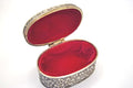 An oval silver-toned keepsake box with an ornate floral engraving pattern on the exterior and a plush red velvet interior, shown open against a plain white background.