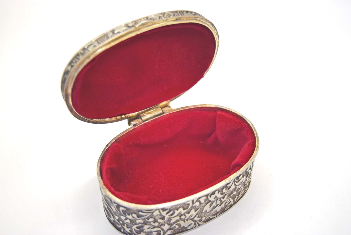 An oval silver-toned keepsake box with an ornate floral engraving pattern on the exterior and a plush red velvet interior, shown open against a plain white background.
