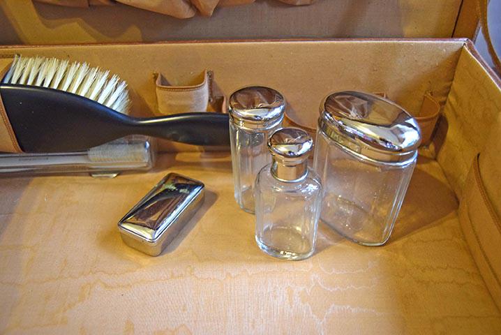 A set of clear glass bathroom or vanity containers with shiny silver metal lids, including two tall cylindrical jars and one shorter bottle, displayed inside a light wood box alongside a black-handled brush and a small silver rectangular item.