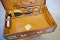 An open vintage brown leather suitcase containing a matching grooming or grooming kit set, including a hairbrush, small metal containers, and a small paper tag, placed on a gray carpeted floor.