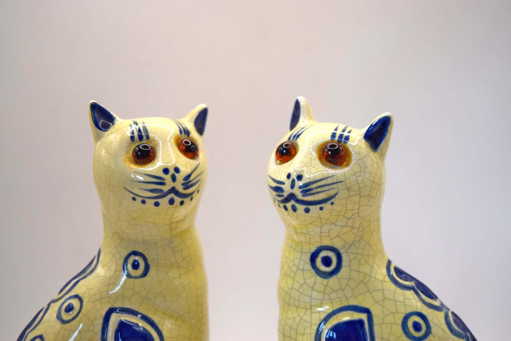 Two ceramic cat figurines with a glossy yellow glaze, blue hand-painted patterns, and realistic brown eyes, set against a plain light background.