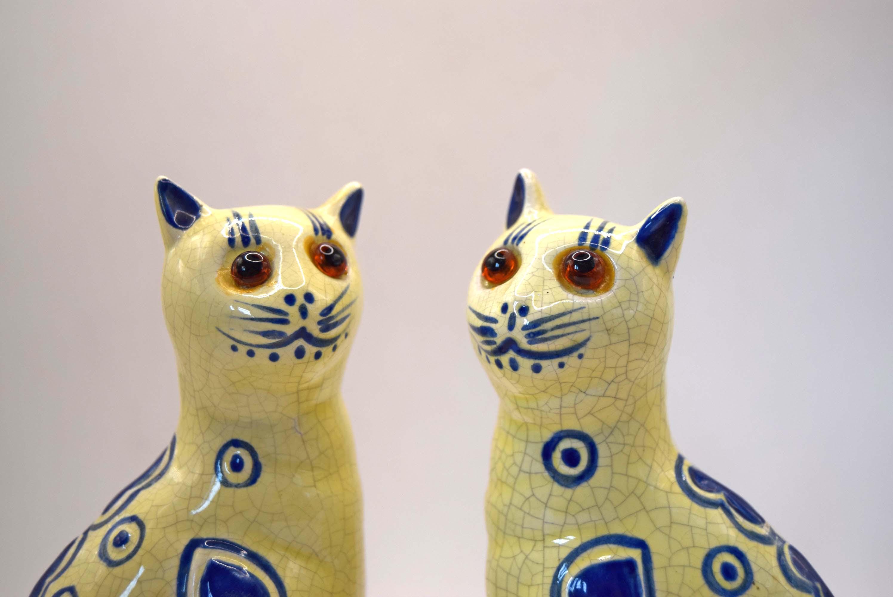Two ceramic cat figurines with a glossy yellow glaze, blue hand-painted patterns, and realistic brown eyes, set against a plain light background.