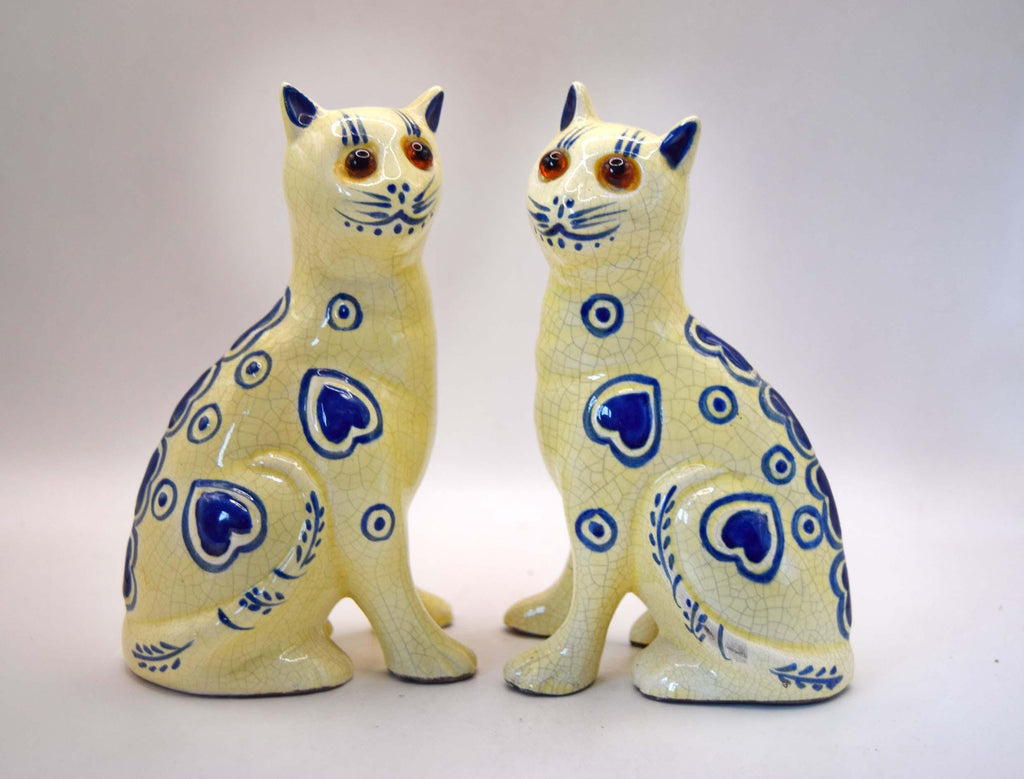 A pair of ceramic cat figurines in a pale cream glaze with blue heart and circle patterns, brown eyes, and blue ears and noses, set against a plain light background.